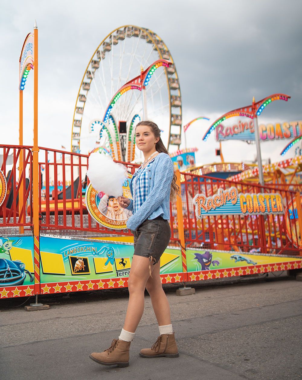 Junge Frau in blau-karierter Trachtenbluse mit hellblauer Trachtenstrickjacke und Ledershorts steht mit Zuckerwatte in der Hand vor einem Riesenrad auf den Cannstatter Wasen