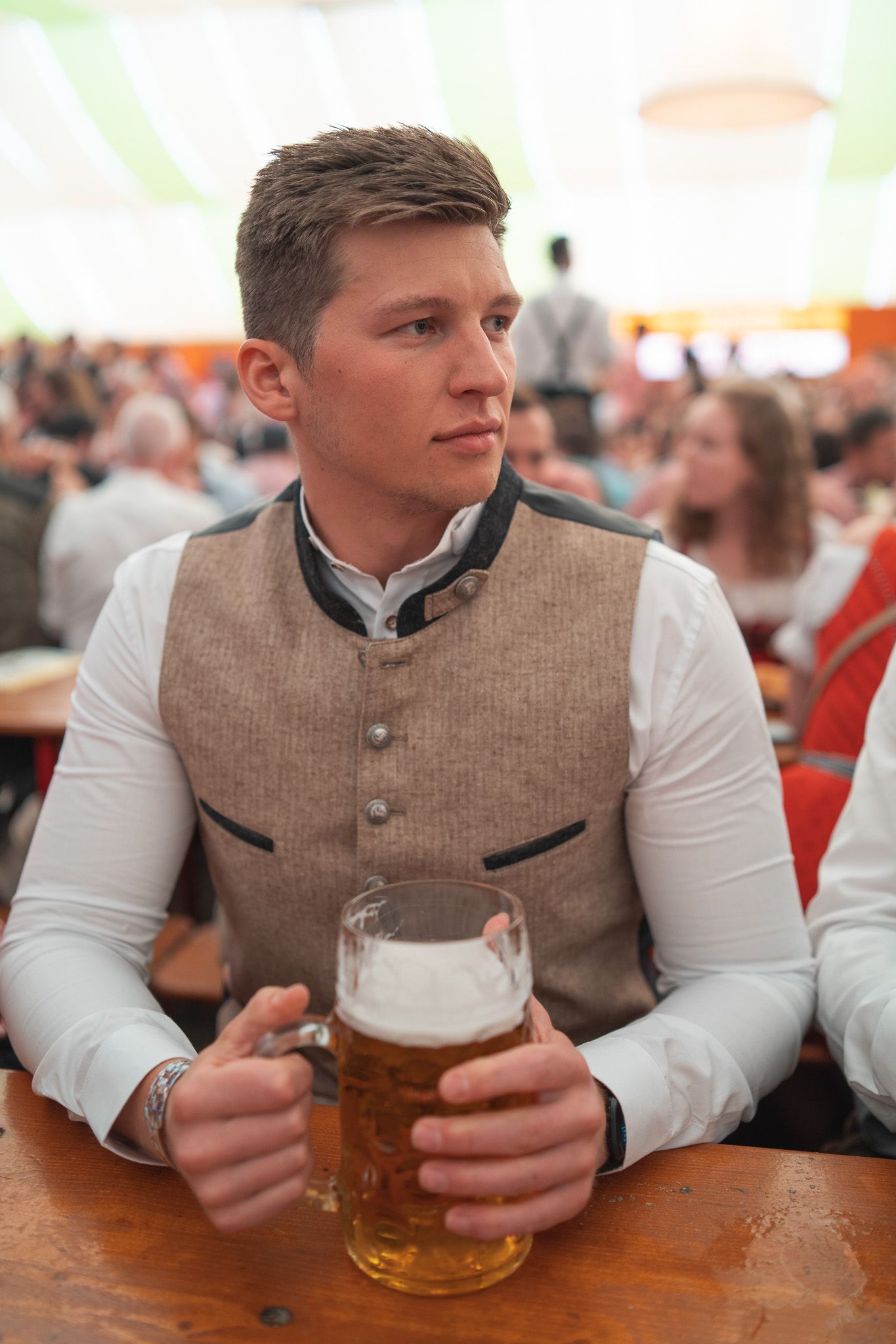 Mann in weißem Trachtenhemd und beige Trachtenweste sitzt mit einem Maßkrug Bier in der Hand in einem Festzelt