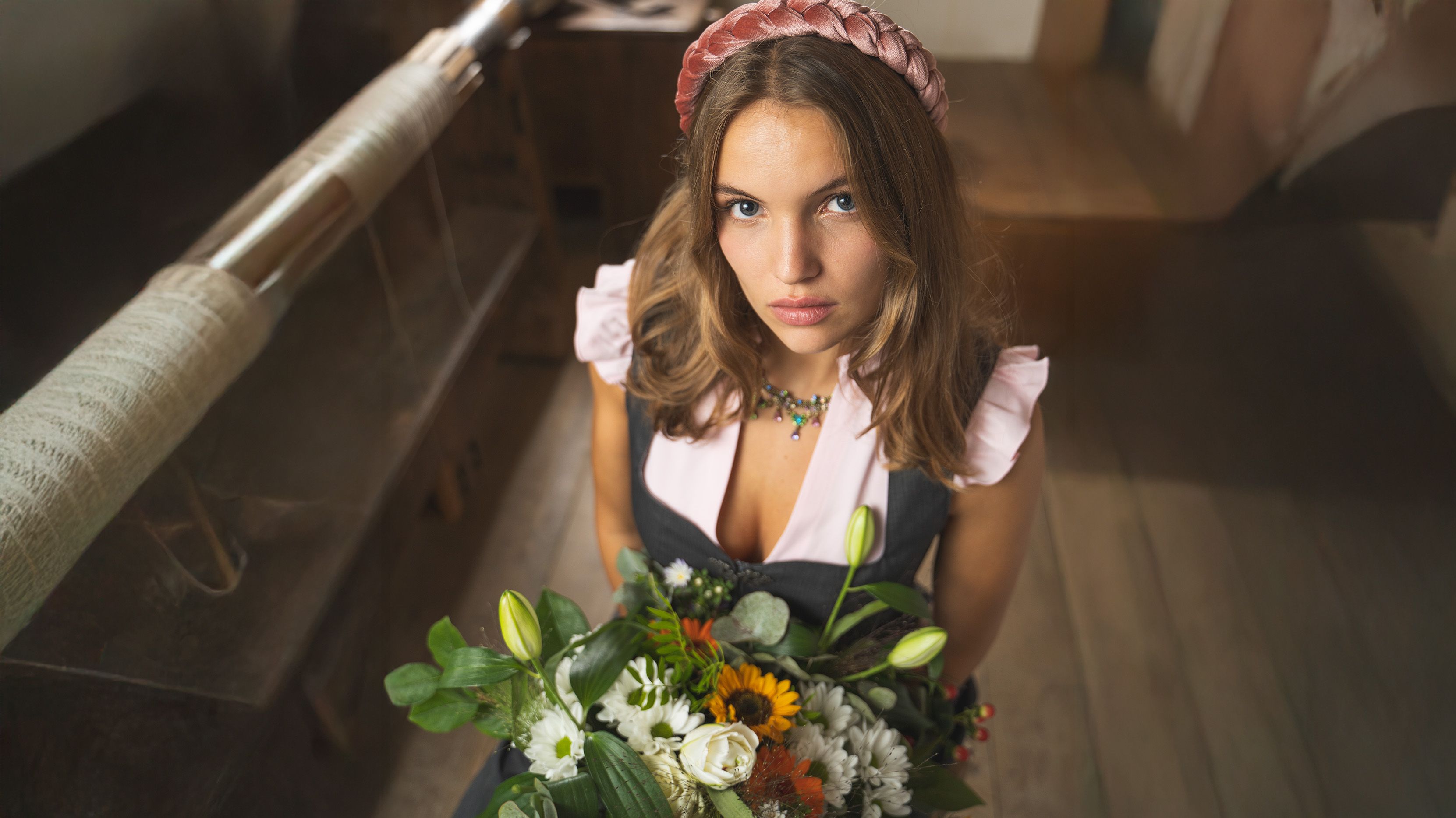 Frau im Dirndl und rosa Flügelbluse sitzt auf einem Holzhocker mit Blumenstrauß in der Hand und guckt von unten nach oben hoch.