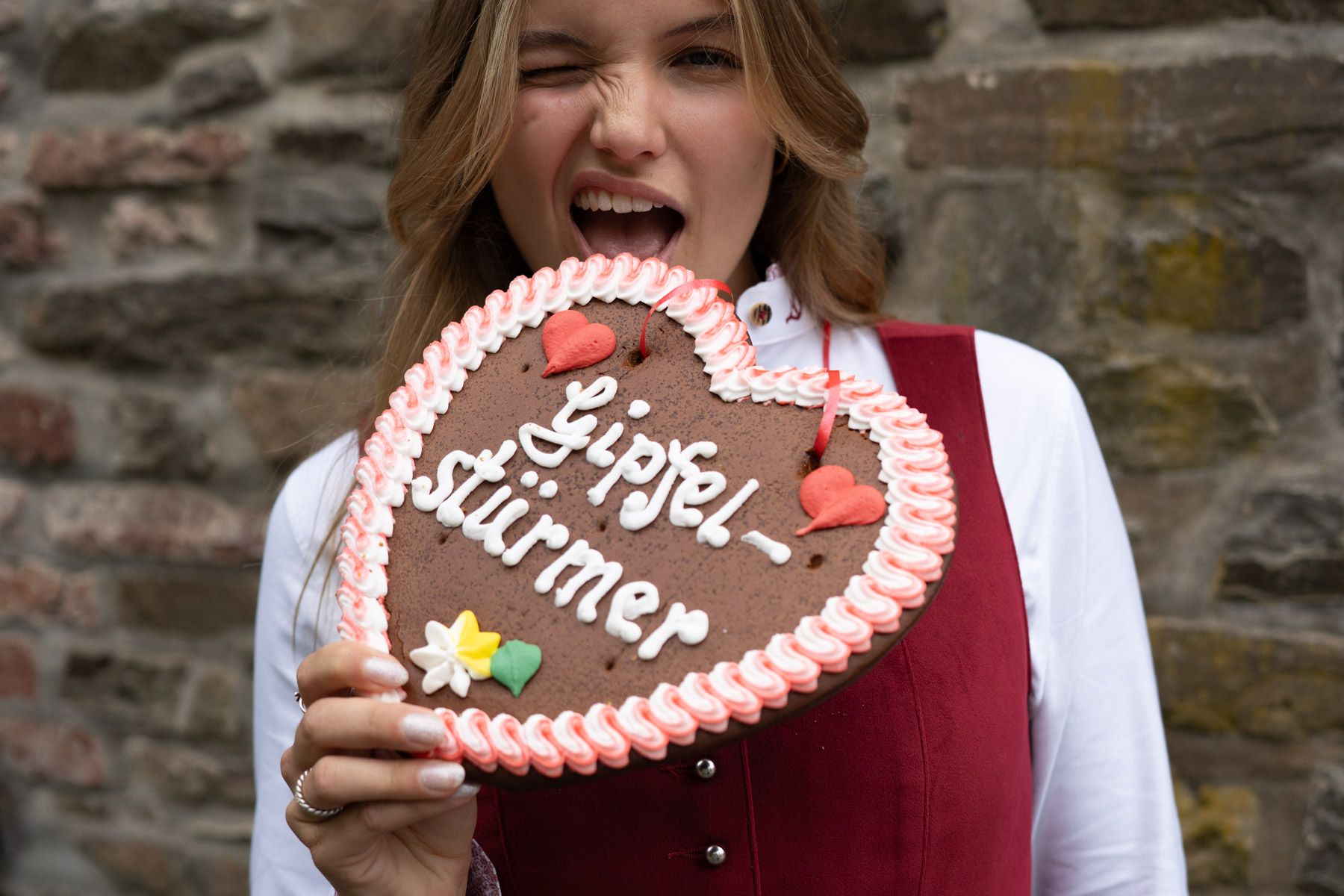 Frau mit rotem Trachtenmieder beißt in ein Lebkuchenherz mit der Aufschrift "Gipfelstürmer".